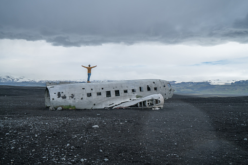 Young man stands arms outstretched on airplane crashed on black sand beach looking around her contemplating surroundings
Famous place to visit in Iceland and pose with the wreck