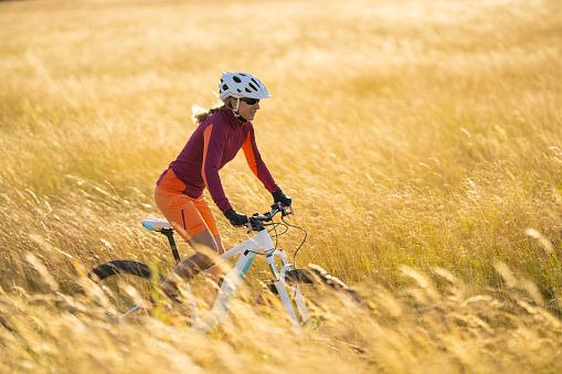 sportive happy 44 years old woman cycling with her electric mountainbike through dry high grass in summer sunlight