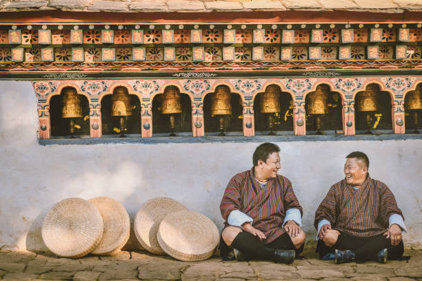 two bhutanese sitting on the ground of temple talk to each other happily two bhutanese sitting on the ground of temple talk to each other happily in front of paro temple , bhutan with the sitting cushion beside them and the prayer wheel wall with their tradition costume on 6th November 2017 buddhist prayer wheel stock pictures, royalty-free photos & images