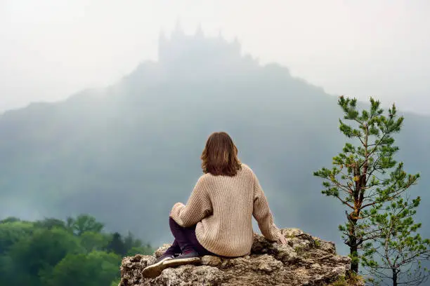 Young female tourist looking on famous Hohenzollern Castle in thick fog, Swabian Alps, Baden-Wurttemberg, Germany. Girl travel around Europe