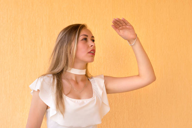 Waist-up portrait of lovely attractive female in white blouse. Blond-haired girl looking far away with hand at her forehead. Isolated on yellow background