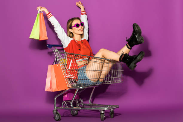 hermosa mujer joven feliz en gafas de sol sosteniendo bolsas de papel y en carro en violeta - adicto a las compras fotografías e imágenes de stock