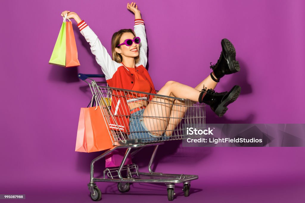beautiful happy young woman in sunglasses holding paper bags and sitting in shopping trolley on violet Shopping Cart Stock Photo
