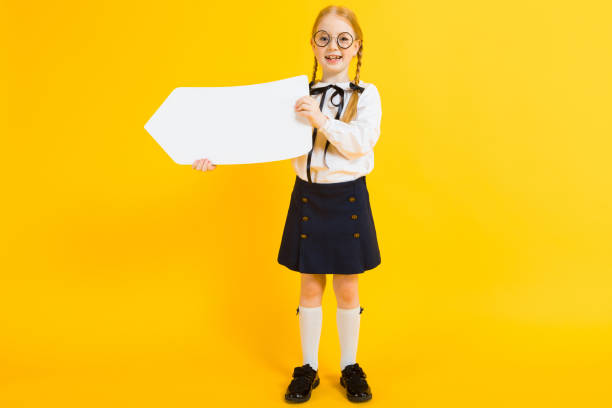 Girl with red pigtails on a yellow background. A charming girl in round transparent glasses is holding a white text box. Portrait of a beautiful girl in a white blouse and black skirt. see through leggings stock pictures, royalty-free photos & images
