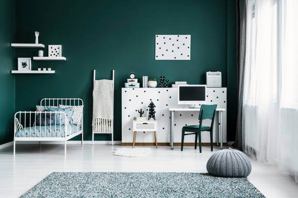 istock Front view of a dark green bedroom interior with a bed, desk, computer, wall decorations and grey pouf on a carpet 995119622