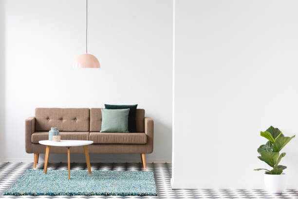 istock Brown sofa, pillows, coffee table and lamp in a living room interior next to an empty wall and plant. Place for your poster or furniture 995119640