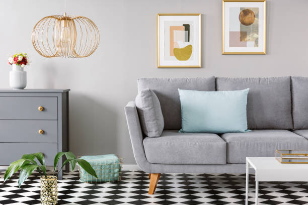 istock Light blue pillow placed on grey couch in bright living room interior with checkerboard linoleum floor, fresh flowers in vase on cupboard two posters hanging on the wall and gold lamp 995141228