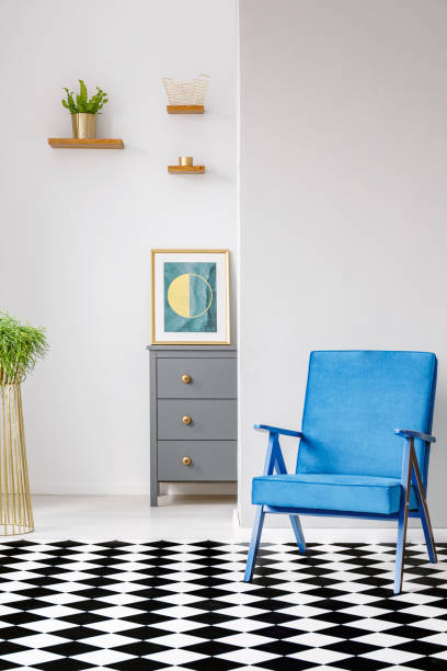 istock Open space room interior with linoleum checkerboard floor, blue armchair, fresh plants and poster standing on grey cupboard 995141234