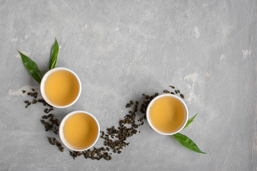 Green tea in ceramic cups, dry green oolong tea and tea leaves on grey stone table, top view, copy space.