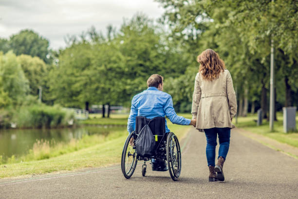 Wheelchair using young man and his girlfriend holding hands whilst walking together through a city park