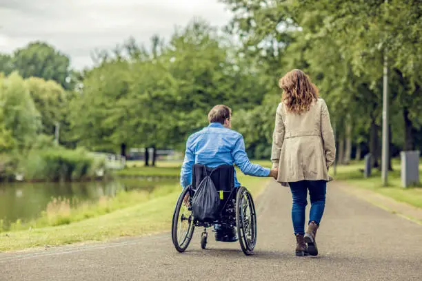 Photo of Wheelchair using young man and his girlfriend holding hands whilst walking together through a city park