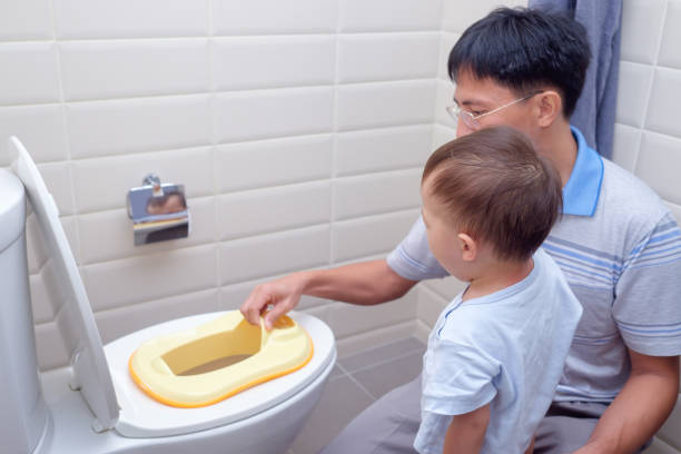 fils somnolent père formation pour utiliser les toilettes dans la salle de bain, mignonne petite asiatique 1 an / bébé de 18 mois pour tout-petits assis sur les toilettes avec accessoire de salle de bain enfant - son photos et images de collection