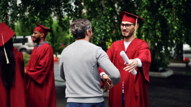 Smiling graduating student is shaking his father's hand and hugging him, young man in glasses is wearing hat and gown and holding diploma. Education and success concept.