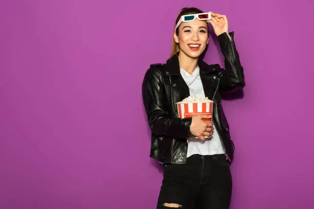 Photo of beautiful happy asian girl in 3d glasses holding popcorn and smiling at camera isolated on violet