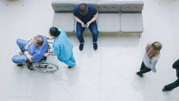 istock High Angle Shot in the Hospital Lobby, Young Man Waits for Results while Sitting and Using Mobile Phone, Doctors, Nurses and Patients Walk Past Him. Clean, New Hospital. 995338548