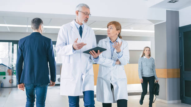 istock In the Hospital, Busy Doctors Talk, Using Tablet Computer While Walking Through the Building. In the Background Patient Talks with Receptionist. New Modern Fully Functional Medical Facility. 995338562