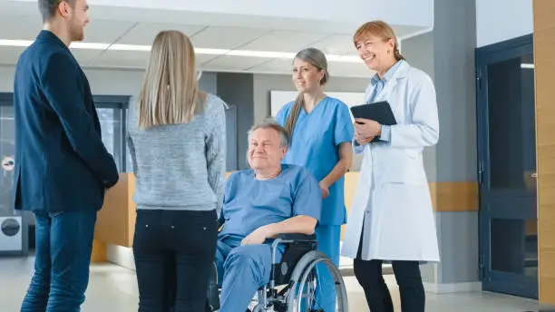 Photo of In the Hospital Lobby. Young Couple Visiting Elderly Parent in a Wheelchair, He's aided by Friendly Nurse and Doctor. Happy Family Reunion. New Modern Medical Facility with Best Possible Personnel.