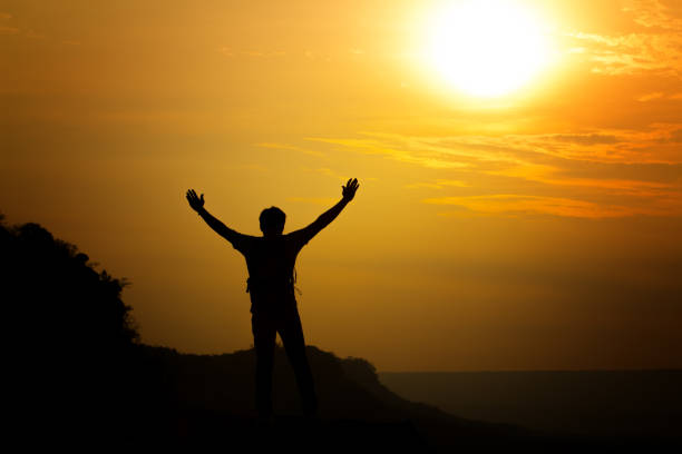istock Silhouette Man Standing With Arms Outstretched Standing On Cliff Against Orange Sky 995368834