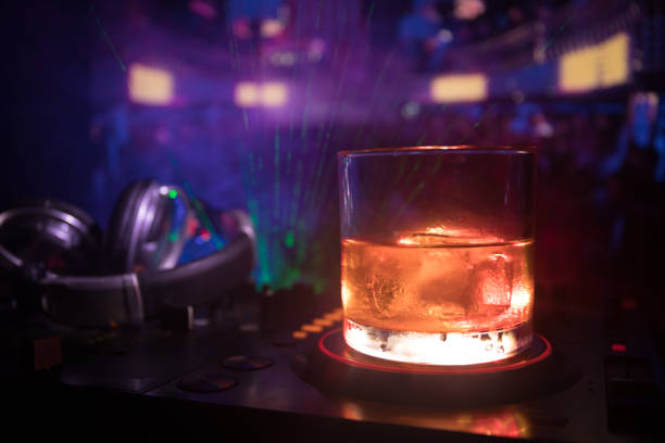 istock Glass with whisky with ice cube inside on dj controller at nightclub. Dj Console with club drink at music party in nightclub with disco lights. 995459816
