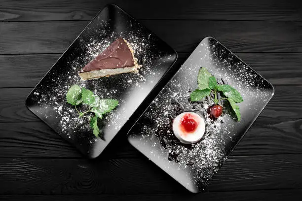 Photo of italian panna cotta dessert with strawberry sirup and cake with nuts and chocolate on the black plate on dark wooden background