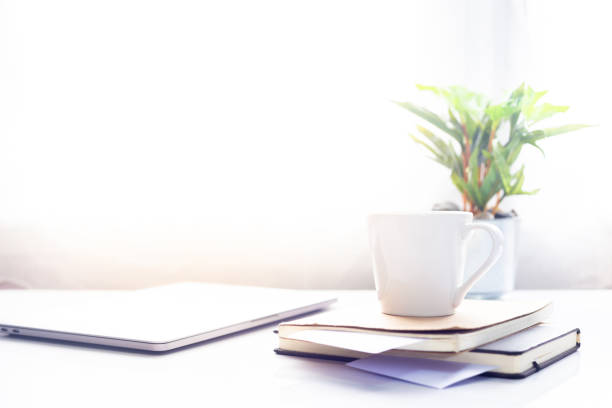 istock Coffee cup on notebook with laptop on white table in the morning sun light ,Working space 995535886
