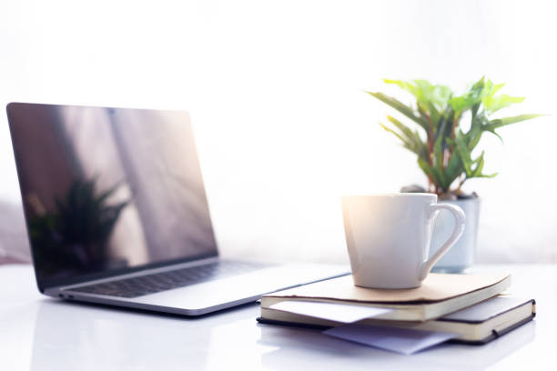 istock Coffee cup on notebook with laptop on white table in the morning sun light ,Working space 995535920