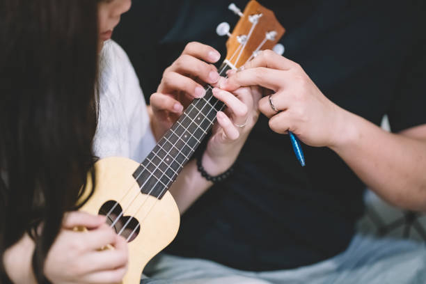 un maschio cinese asiatico insegnamento di una femmina cinese asiatica su ukulele in soggiorno - plucking an instrument foto e immagini stock