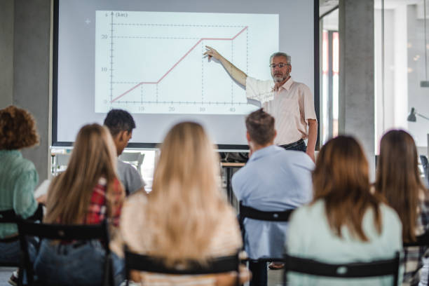 Mature professor giving a presentation to large group of students. Mature teacher teaching group of university students while aiming at projection screen at lecture hall. teacher projector stock pictures, royalty-free photos & images