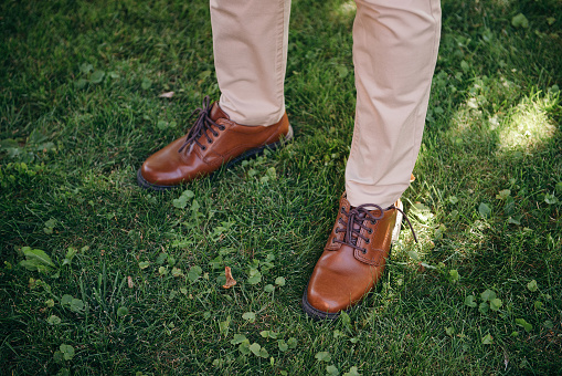 Brown leather shoes on green grass. Man's leg wearing elegant, smart pants and shoes standing in summer park in sunny day