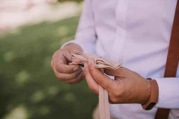 Man holding stylish bow tie in hands. Guy wearing shirt and suspenders is going to fix bow tie.  Trendy groom, fiance outside in green nature