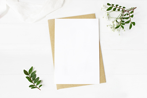 Feminine wedding stationery, desktop mock-up scene. Blank greeting card, craft envelope, baby's breath flowers, silk ribbon and lentisk branches, old white wooden table background. Flat lay, top view.