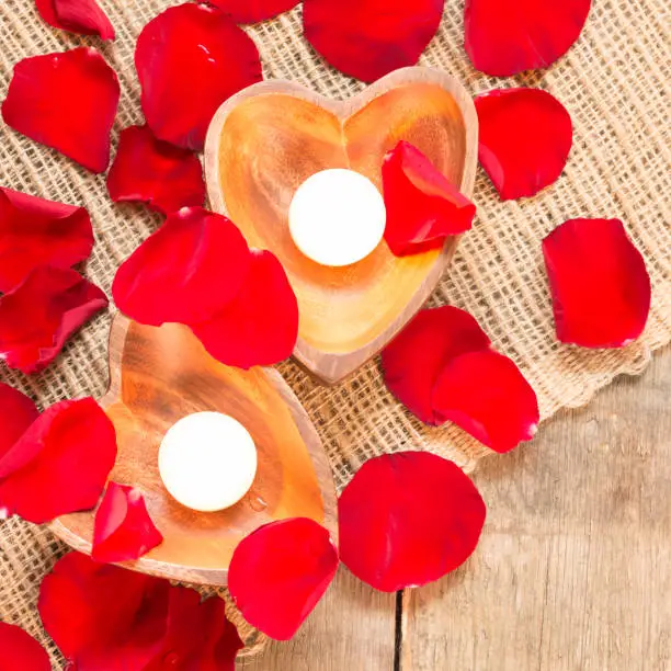 Enlightened candles in heart-shaped candleholders with red roses petals on rustic wooden background. St Valentines background. Romantic holiday concept. Top view. Square