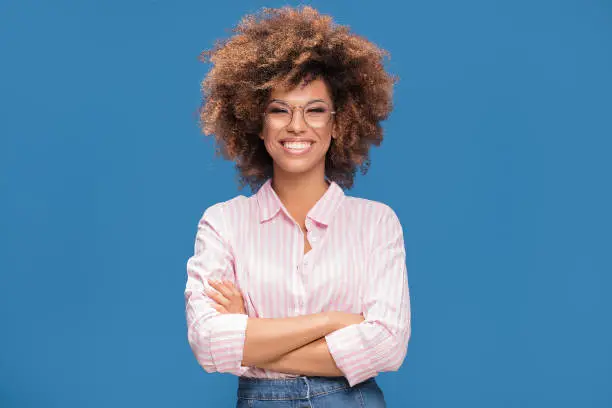 Closeup portrait of Afro American woman with bushy hair wearing fashionable eyeglasses ,looking at camera and smiling, blue background.