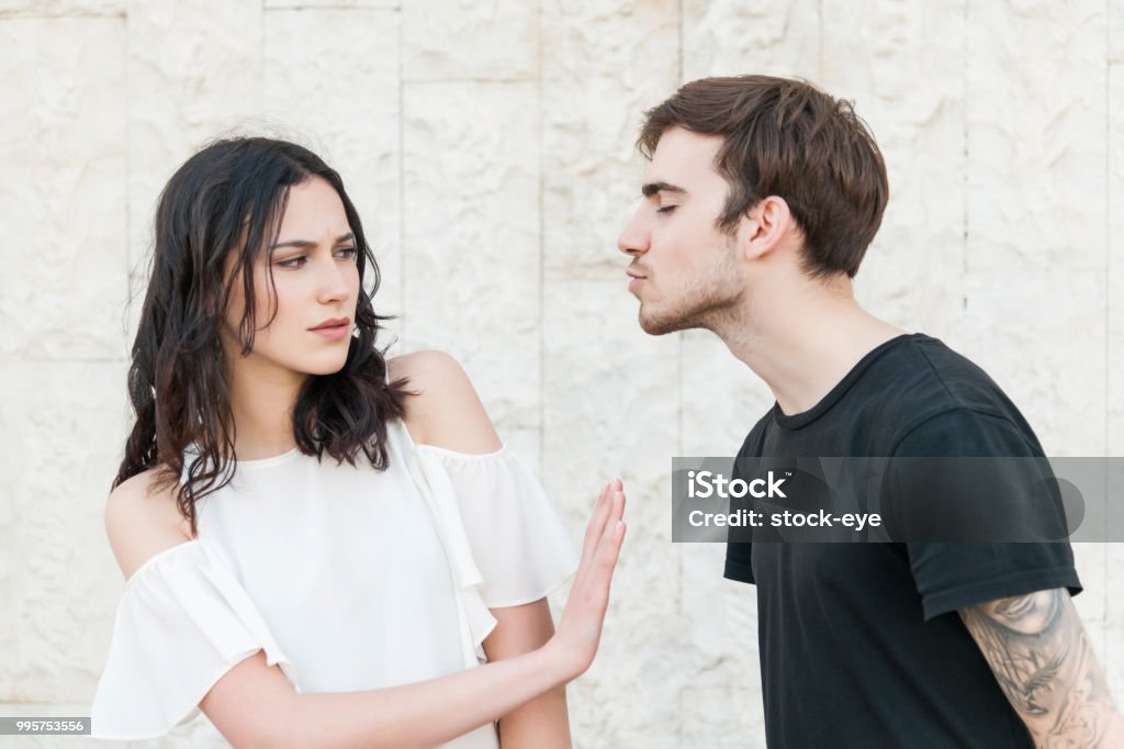 Young man trying to kiss a young woman A young guy is leaning in to kiss a girl be she is frowning and making a hand gesture to stop him. They are wearing casual clothes. Rejection Stock Photo