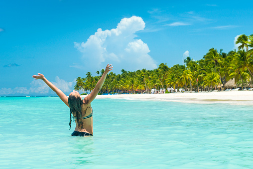 Back view of a happy teenage girl with her arms wide open in the Caribbean ocean. She is standing at the beach of the island Saona in the Dominican Republic.