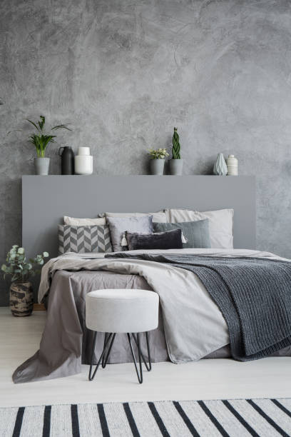 istock Striped carpet and stool in grey bedroom interior with sheets on bed with headboard. Real photo 995800012