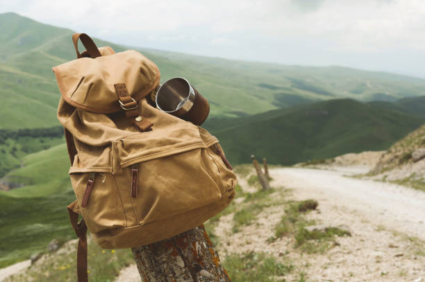 istock Hipster yellow vintage backpack with a mug fixed on it with a mug close-up front view. Traveler's travel bag in the background of a mountain landscape and a road 995929672