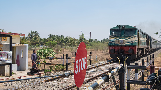 Tiruppur, India - March 8, 2018: A passenger train on the southern section of Indian railways approaching a manned road crossing.\nThe national rail network extends over 75,000 miles of track and transports 23 million passengers each day