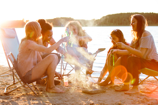 small group of young women enjoy a carefree summer day at a lake while making marshmallows over a campfire