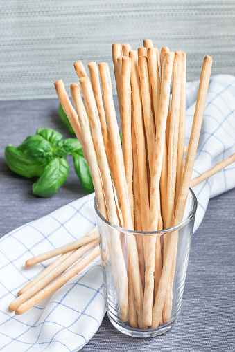Traditional Italian snack grissini bread sticks in glass with basil leaves on background, vertical