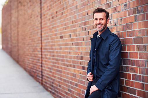 Smiling middle-aged businessman staring at camera and leaning on brick wall