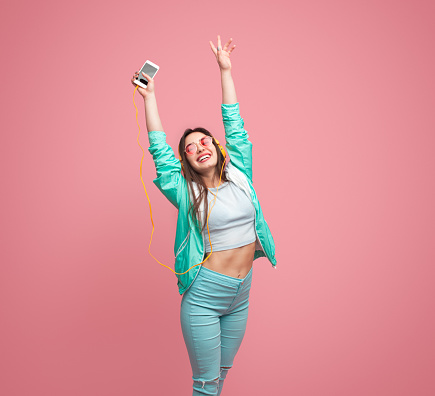 Stylish smiling female in casual outfit holding modern smartphone while standing with hands up and listening music through big yellow earphones on pink background