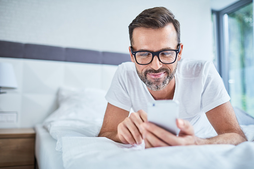 Happy man lying on bed and using phone in the morning
