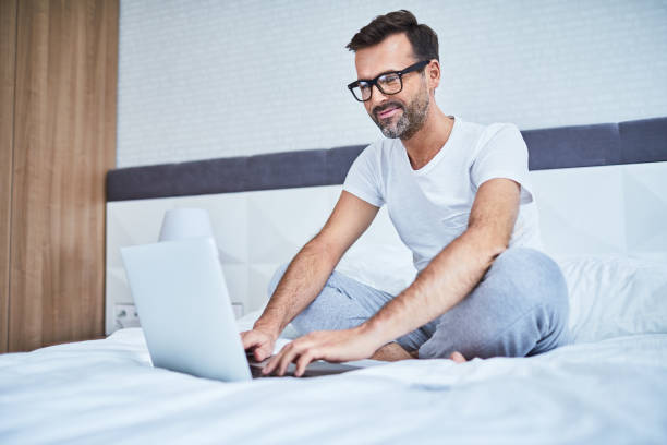 istock Smiling man sitting on bed and typing on laptop 996087916