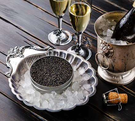 Black caviar in can on ice in silver bowl and champagne on black wooden background