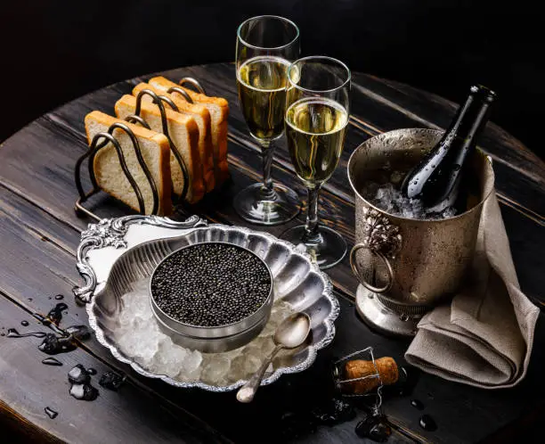 Black caviar in can on ice in silver bowl, bread and champagne in ice bucket on black wooden background