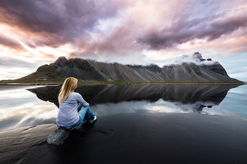 Rear view of woman sitting on a rock at the beach and looking at majestic  Vestrahorn mountain in Iceland. Copy space.
