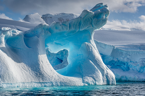 A dramatically shaped and weather eroded iceberg floating in Wilhemina Bay Antarctica with a large hole in the centre glowing blue, and with a distant background of snow and ice covered mountains on a a sunlit summers day