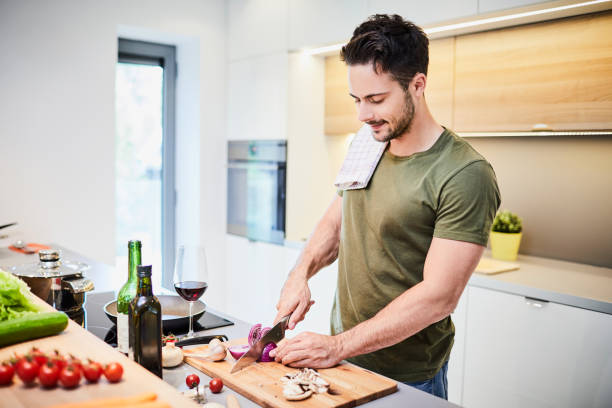 bonito jovem preparando jantar helathy, em pé no fogão e legumes corte - helathy - fotografias e filmes do acervo