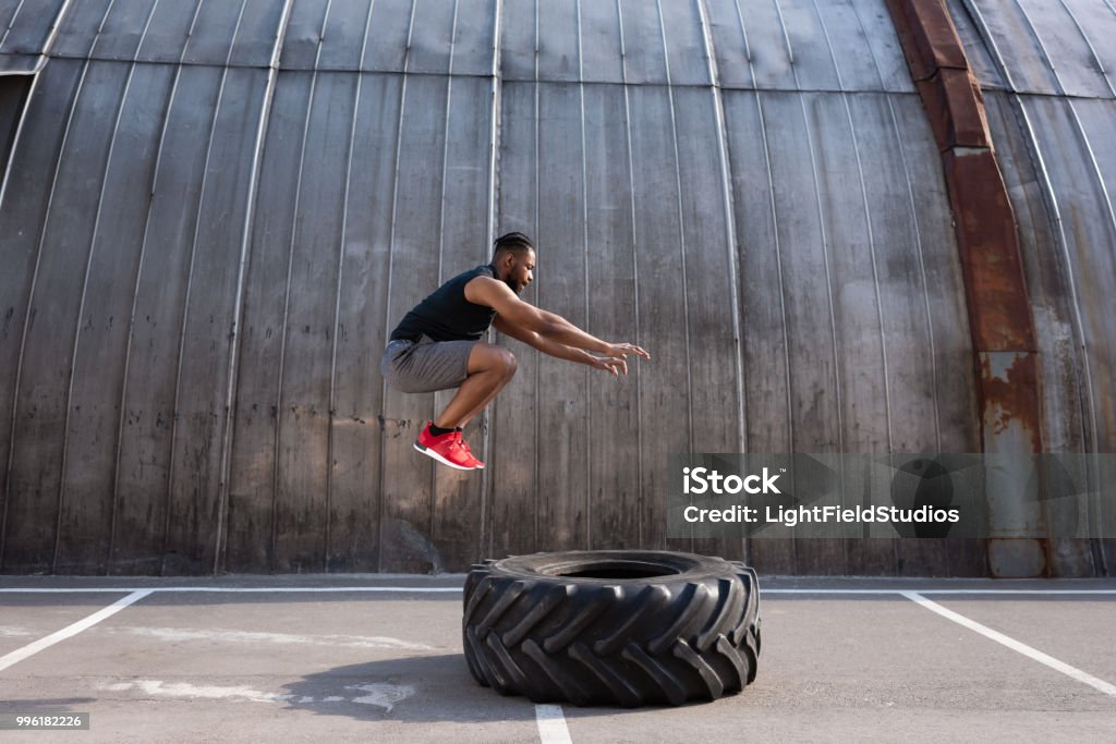 muscular african american sportsman jumping while training with tyre on street Exercising Stock Photo